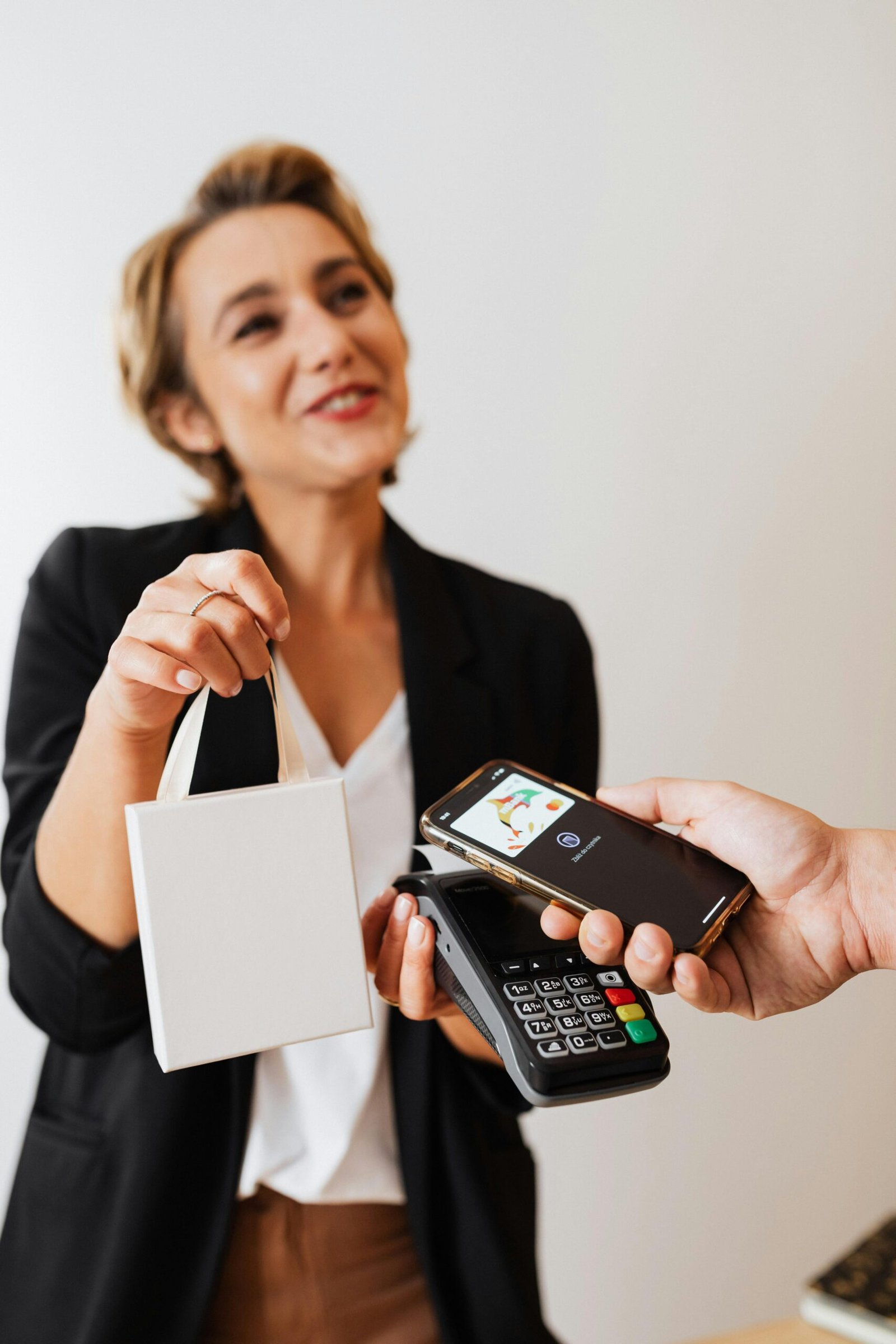 Woman using mobile phone for contactless payment at a cash register with a small shopping bag.